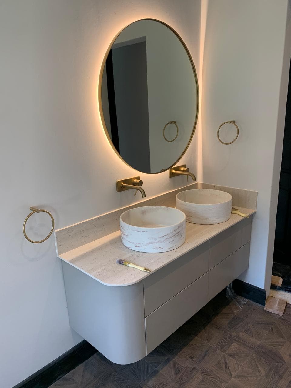 A curved, floating vanity in soft stone grey with two round basins under a back-lit brass mirror