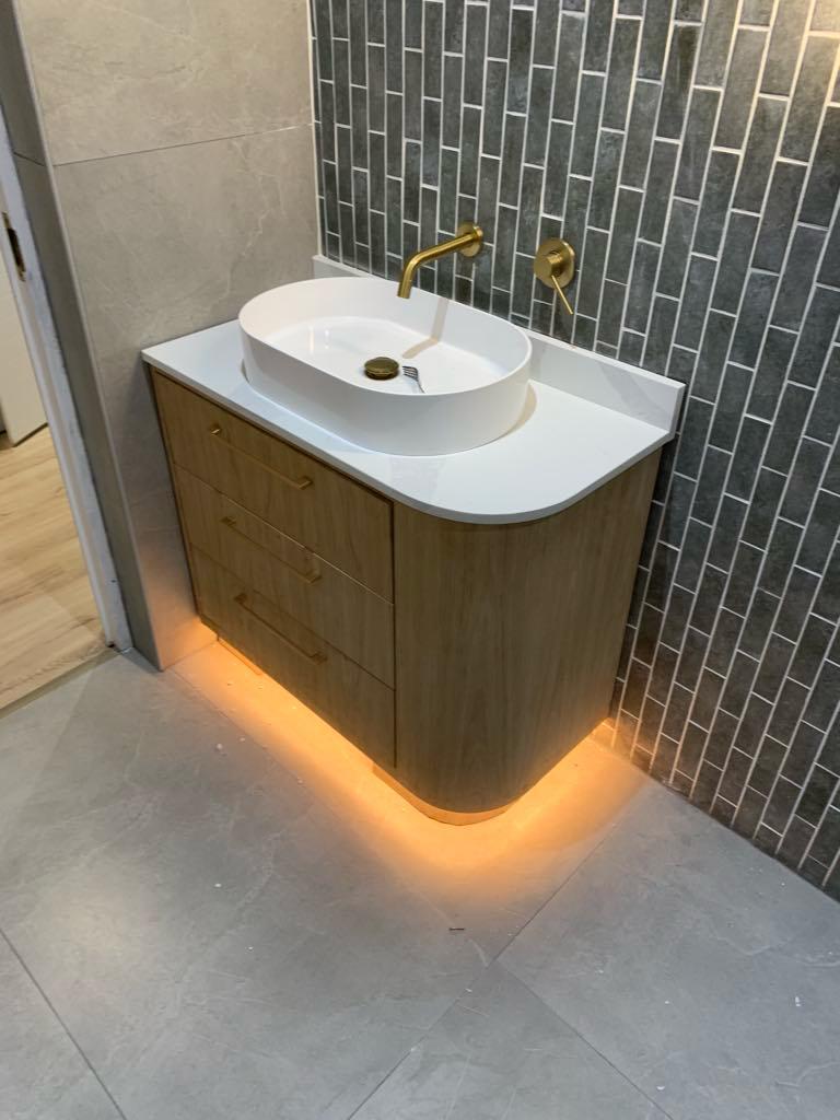 Curved oak vanity with an oval basin, brass taps and warm under-lighting